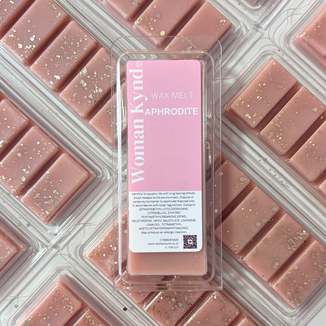 Packaged wax melts with a pink label, named 'APHRODITE', by Woman by Design. The package contains rectangular wax pieces visible through a clear plastic container, with gold flecks within the wax.