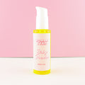 A bottle of Woman Kynd Baby Powder Body Oil with a white pump dispenser and a white label, filled with body oil, set against a pink and white background.