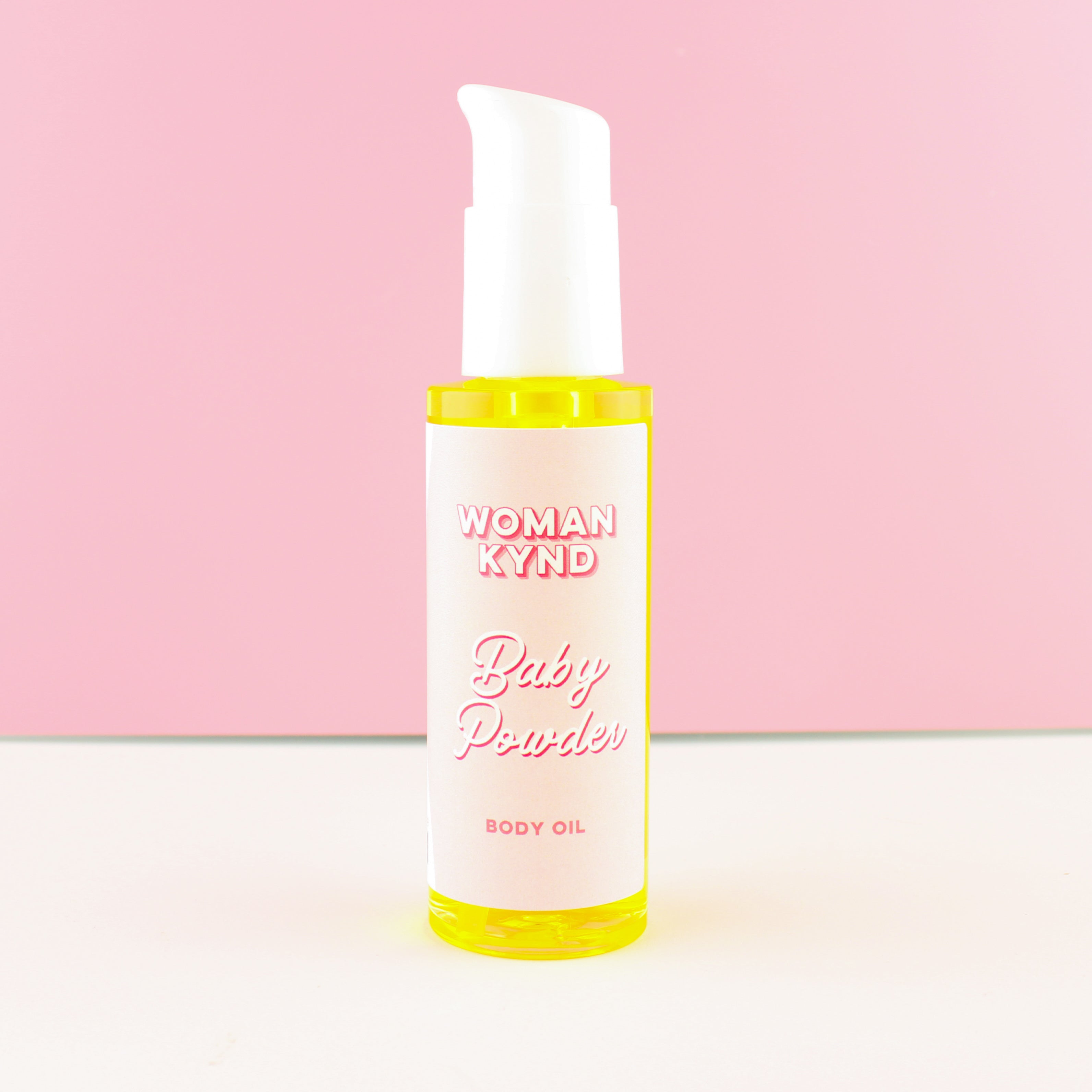 A bottle of Woman Kynd Baby Powder Body Oil with a white pump dispenser and a white label, filled with body oil, set against a pink and white background.