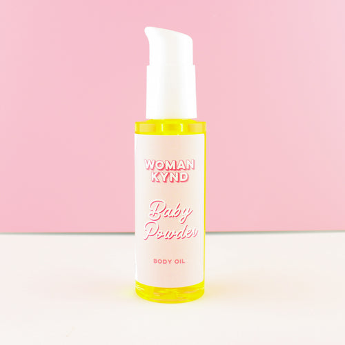 A bottle of Woman Kynd Baby Powder Body Oil with a white pump dispenser and a white label, filled with body oil, set against a pink and white background.