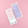 Two images of a Woman Kynd five segment light purple Wax Melt with dark purple glitter, both in a frosted clamshell. The first image displays the pink and white label with the words Woman Kynd Black Cherry Wax Melt and the CLP information, and the other image is of the reverse side showing the Wax Melt Itself. Both images are against a solid pink background.