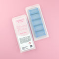 Two images of a Woman Kynd five segment light blue Wax Melt with dark blue glitter, both in a frosted clamshell. The first image displays the pink and white label with the words Woman Kynd Blueberry Frosting Wax Melt and the CLP information, and the other image is of the reverse side showing the Wax Melt Itself. Both images are against a solid pink background.