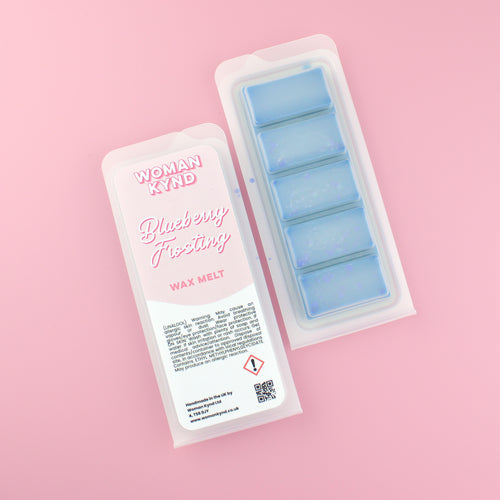 Two images of a Woman Kynd five segment light blue Wax Melt with dark blue glitter, both in a frosted clamshell. The first image displays the pink and white label with the words Woman Kynd Blueberry Frosting Wax Melt and the CLP information, and the other image is of the reverse side showing the Wax Melt Itself. Both images are against a solid pink background.