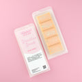 Two images of a Woman Kynd five segment orange Wax Melt with gold glitter, both in a frosted clamshell. The first image displays the pink and white label with the words Woman Kynd Brazilian Beach Wax Melt and the CLP information, and the other image is of the reverse side showing the Wax Melt Itself. Both images are against a solid pink background.