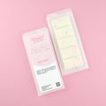 Two images of a Woman Kynd five segment beige Wax Melt with pink and blue glitter, both in a frosted clamshell. The first image displays the pink and white label with the words Woman Kynd Cherry Blossom & Nordic Cotton Wax Melt and the CLP information, and the other image is of the reverse side showing the Wax Melt Itself. Both images are against a solid pink background.