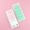 Two images of a Woman Kynd five segment mint green Wax Melt with white and gold glitter, both in a frosted clamshell. The first image displays the pink and white label with the words Woman Kynd Coco Cabana Wax Melt and the CLP information, and the other image is of the reverse side showing the Wax Melt Itself. Both images are against a solid pink background.