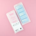 Two images of a Woman Kynd five segment light blue Wax Melt with white wax patterns in the shape of clouds, both in a frosted clamshell. The first image displays the pink and white label with the words Woman Kynd Fuffy Skies Wax Melt and the CLP information, and the other image is of the reverse side showing the Wax Melt Itself. Both images are against a solid pink background.