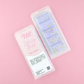 Two images of a Woman Kynd five segment light purple Wax Melt with dark purple and yellow glitter, both in a frosted clamshell. The first image displays the pink and white label with the words Woman Kynd Lullaby Baby Wax Melt and the CLP information, and the other image is of the reverse side showing the Wax Melt Itself. Both images are against a solid pink background.