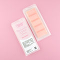 Two images of a Woman Kynd five segment pale orange Wax Melt with pale gold glitter, both in a frosted clamshell. The first image displays the pink and white label with the words Woman Kynd Marshmallow & White Peach Wax Melt and the CLP information, and the other image is of the reverse side showing the Wax Melt Itself. Both images are against a solid pink background.