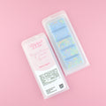 Two images of a Woman Kynd five segment light blue Wax Melt with yellow and green glitter, both in a frosted clamshell. The first image displays the pink and white label with the words Woman Kynd Orange Blossom & Coastal Cypress Wax Melt and the CLP information, and the other image is of the reverse side showing the Wax Melt Itself. Both images are against a solid pink background.