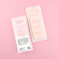 Two images of a Woman Kynd five segment pale pink Wax Melt with pink glitter, both in a frosted clamshell. The first image displays the pink and white label with the words Woman Kynd Pink Vanilla & Coco Blossom Wax Melt and the CLP information, and the other image is of the reverse side showing the Wax Melt Itself. Both images are against a solid pink background.