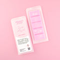 Two images of a Woman Kynd five segment pink Wax Melt with white, snowflake shaped glitter, both in a frosted clamshell. The first image displays the pink and white label with the words Woman Kynd Snow Angel Wax Melt and the CLP information, and the other image is of the reverse side showing the Wax Melt Itself. Both images are against a solid pink background.