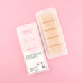 Two images of a Woman Kynd five segment beige Wax Melt with gold glitter, both in a frosted clamshell. The first image displays the pink and white label with the words Woman Kynd Spa Day Wax Melt and the CLP information, and the other image is of the reverse side showing the Wax Melt Itself. Both images are against a solid pink background.
