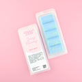 Two images of a Woman Kynd five segment light blue Wax Melt with green glitter, both in a frosted clamshell. The first image displays the pink and white label with the words Woman Kynd Spring Morning Wax Melt and the CLP information, and the other image is of the reverse side showing the Wax Melt Itself. Both images are against a solid pink background.